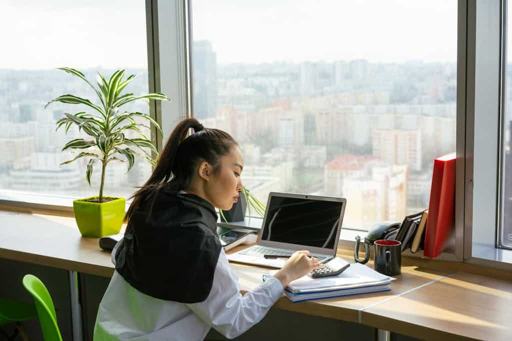Asian woman focusing on work in a modern high-rise office setting with city view.